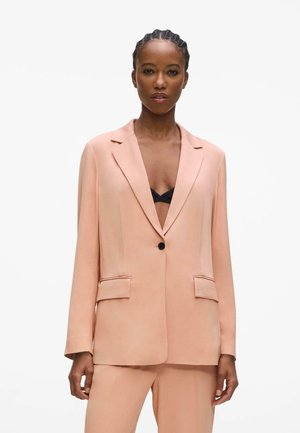 Kobieta z warkoczami, ubrana w brzoskwiniową marynarkę i pasujące spodnie na czarnym biustonoszu, stojąca na białym tle.