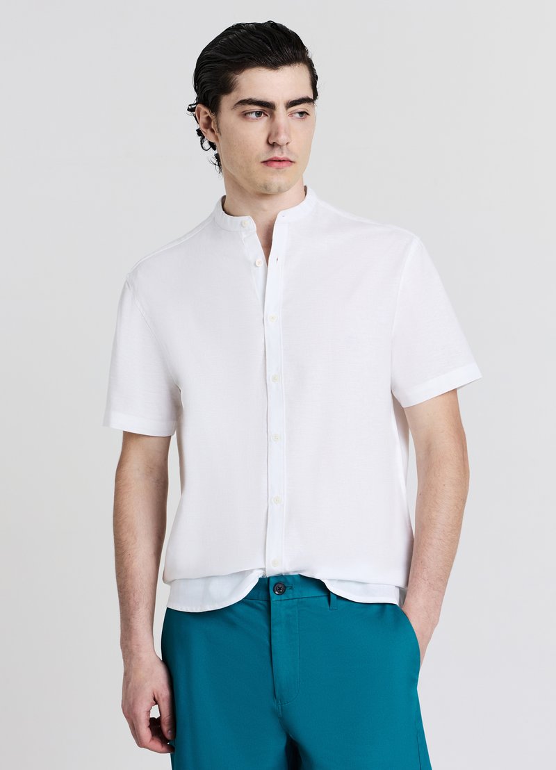 Chemise blanche à manches courtes avec col montant et fermeture par boutons, en tissu léger, associée à un pantalon bleu sarcelle. Design minimaliste.