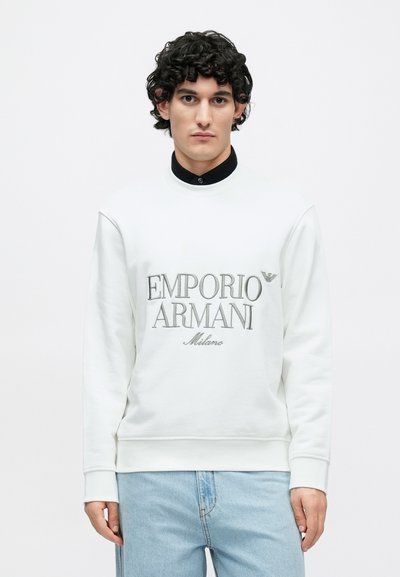 Sweatshirt branca com logótipo cinzento "Emporio Armani Milano", textura suave, gola redonda e punhos canelados. Combinada com jeans azul claro.