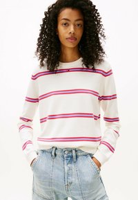 Femme aux cheveux bouclés portant un pull blanc avec des rayures violettes et rouges, un jean taille haute bleu clair, les mains dans les poches, regardant droit devant.