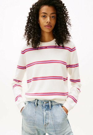 Frau mit lockigem Haar, die einen weißen Pullover mit lila und roten Streifen sowie hellblauen, hoch taillierten Jeans trägt, mit den Händen in den Taschen und nach vorne blickend.