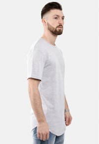 Graues T-Shirt mit kurzen Ärmeln und rundem Halsausschnitt. Weicher, leichter Stoff. Leicht verlängerter Saum hinten. Kombiniert mit abgetragenen Jeans.