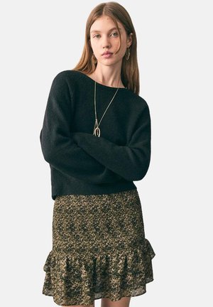 Une jeune femme aux cheveux longs porte un pull noir, une jupe à volants à motifs, un collier en or et des boucles d'oreilles, se tenant les bras croisés.