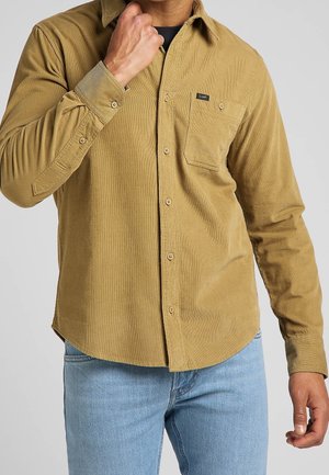 Uomo che indossa una camicia color crema in velluto a coste con taschino sul petto e jeans azzurri, mano vicino al colletto, su uno sfondo grigio uniforme.