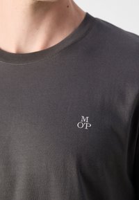 Gros plan sur une personne portant un t-shirt à col rond gris foncé avec un petit logo blanc "M O P" sur le côté gauche de la poitrine.