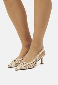Un paio di tacchi slingback open-toe in oro metallico, caratterizzati da un design in rete intrecciata e un tacco kitten, che mostrano una forma a punta.