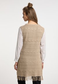 Gilet lungo in crochet beige con motivi texture e dettagli con frange all'orlo, indossato su una camicia bianca a maniche lunghe.