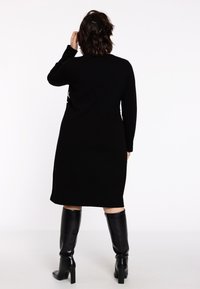 Une femme aux cheveux sombres mi-longs, portant une robe noire et des bottes noires jusqu'aux genoux, se tient dos au spectateur sur un fond blanc.