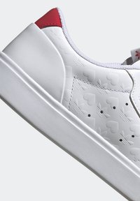 Vit sneaker med präglat hjärta och logotypmönster, röda detaljer på hälen, slät läderyta och en solid vit gummisula.