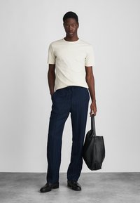 Κρέμα κοντομάνικη μπλούζα συνδυασμένη με σκούρα μπλε παντελόνι με υφή και ρίγες. Το μοντέλο κρατάει μια μαύρη δερμάτινη τσάντα και στέκεται σε μια γκρίζα επιφάνεια.