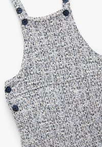 Salopette in tweed blu e bianco, con spalline regolabili, caratterizzata da grandi bottoni scuri e un motivo testurizzato su tutta la superficie.