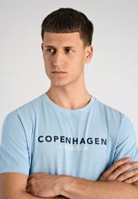 Maglietta a maniche corte di colore azzurro realizzata in tessuto morbido, con la scritta "COPENHAGEN LINDBERGH" in caratteri blu scuro e bianchi.