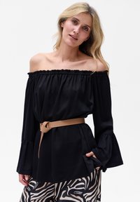 Schwarze Off-Shoulder-Bluse mit elastischem Halsausschnitt, langen Glockenärmeln und tailliertem Design. Kombiniert mit gemusterten beigen und schwarzen Hosen.