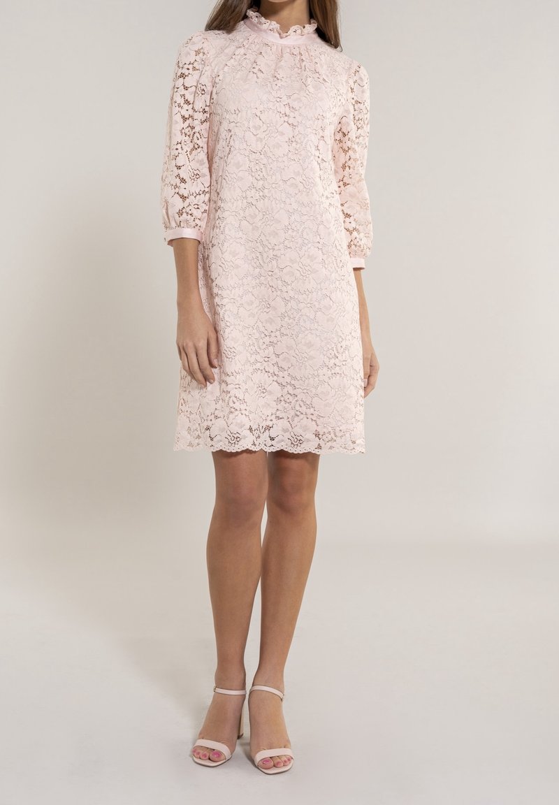 Femme portant une robe en dentelle rose pâle mi-longue avec des manches trois-quarts et un col montant à volants, associée à des talons ouverts assortis.