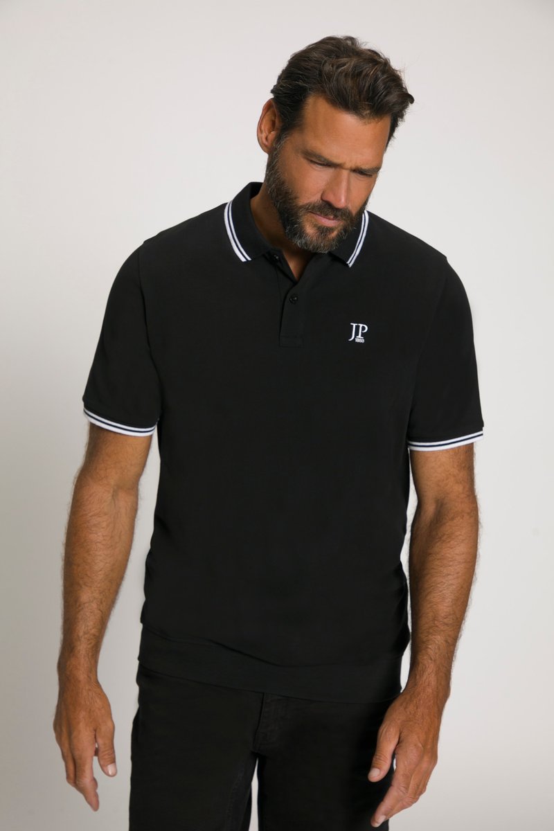 Bärtiger Mann, der ein schwarzes Poloshirt mit weißem Besatz am Kragen und an den Ärmeln trägt, schaut nach unten, steht vor einem schlichten hellen Hintergrund.
