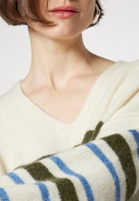 Sweter w serek z kremowej wełny z zielonymi i niebieskimi paskami na rękawach; miękka tekstura, luźny krój i widoczne detale szwów.