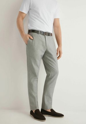 Mężczyzna ubrany w jasnoszare spodnie, białą koszulę z krótkim rękawem, ciemny pasek i ciemnobrązowe buty wsuwane, stojący z jedną ręką w kieszeni.