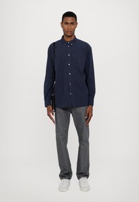 Marineblauw verticaal gestreept overhemd met een borstzak, gecombineerd met donkergrijze jeans en witte sneakers. Het overhemd heeft lange mouwen.