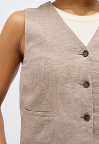 Αμάνικο μπεζ γιλέκο με τρία σκουρόχρωμα κουμπιά πάνω από λευκή ριπ αμάνικη μπλούζα σε άτομο με μέτριο τόνο δέρματος.