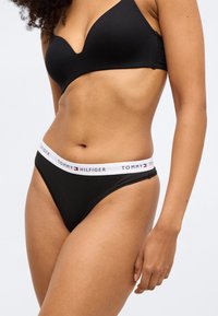 Ženska, ki nosi črni Tommy Hilfiger bralette in ujemajoče črne spodnje perilo z belim pasom z blagovno znamko, pozira na belem ozadju.