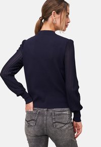 Pull en bleu marine avec un dos lisse et des manches longues plissées texturées. Présente des poignets côtelés. Assorti à un jean gris clair.