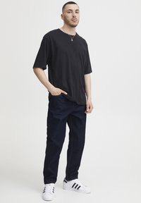 Camiseta negra de cuello redondo, ajuste relajado. Vaqueros de mezclilla azul oscuro. Zapatillas blancas con rayas negras. Collar de oro como acento. Atuendo simple y casual.