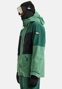 Groene en zwarte waterdichte jas met een capuchon, ritssluiting zakken en een logopatch. Textuurstof, ontworpen voor buitengebruik.