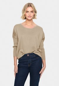 Maglione beige lavorato a maglia con scollatura ampia e maniche a tre quarti, abbinato a jeans blu scuro con tasca e cuciture a contrasto.