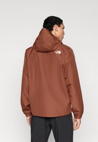 Καφέ αδιάβροχο μπουφάν με κουκούλα, ελαστικά μανίκια και μικρό λευκό λογότυπο στην πλάτη. Απλός, λειτουργικός σχεδιασμός, κατάλληλος για υπαίθρια χρήση.