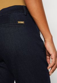 Pantalones de mezclilla de un azul oscuro con un bolsillo trasero que tiene una etiqueta de metal de la marca, costuras visibles y una textura suave.