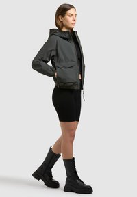 Woman standing, wearing a dark green hooded jacket, black cropped shorts, and black lace-up combat boots against a plain background.