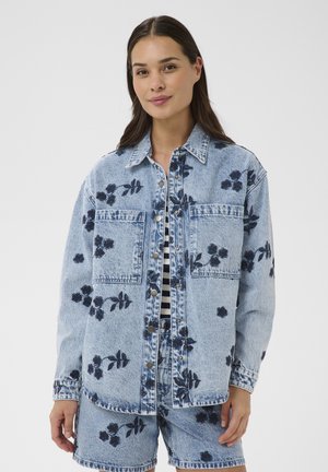 Femme portant une veste en jean bleu clair et un short assorti avec des imprimés floraux bleu foncé, debout devant un fond blanc uni.