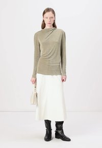 Olive green long-sleeve top with draped design, paired with a white midi skirt and black knee-high boots. Holding a small beige handbag.