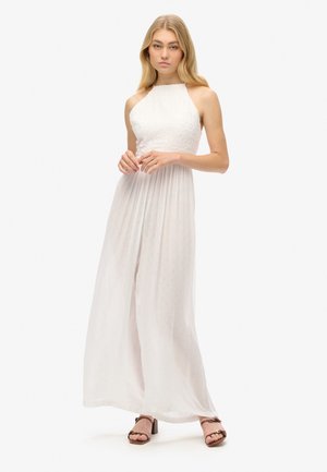 Robe maxi blanche à encolure halter avec un bustier texturé, une jupe plissée et un tissu léger. Le mannequin porte des sandales à talons carrés marron.
