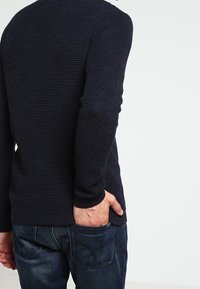 Pull texturé bleu marine en maille avec manches longues, col rond et poignets côtelés, associé à un jean en denim foncé. Main placée dans la poche.