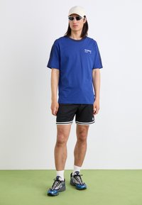 T-shirt en coton bleu avec des manches noires, shorts en mesh noirs avec des accents blancs, et chaussures de sport bleues. Accompagné d'une casquette beige et de lunettes de soleil.