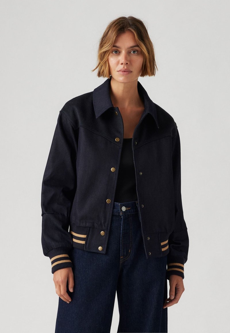 Veste en denim foncé avec un devant à boutons, un col pointu et des poignets côtelés avec des rayures dorées. Texture lisse, coupe décontractée.