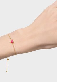 Pulsera de cadena de oro con una gema rosa en forma de corazón engastada en un soporte. Presenta un regulador deslizante y extremos de cadena colgantes.