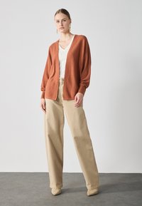 Rustrød, oversized cardigan med lange ærmer, båret over en hvid V-hals top og beige vide bukser. Blød tekstur og løs pasform.