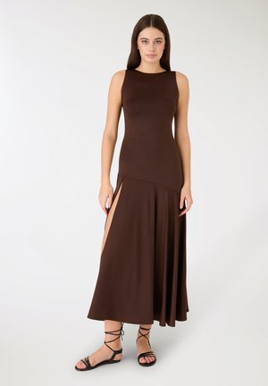 Abito lungo marrone senza maniche con corpetto aderente e gonna fluente, caratterizzato da spacchi laterali. Il materiale sembra liscio e morbido. Sandali neri con cinturini.