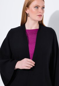 Schwarze Oberbekleidung mit einem Schalkragen und weiten Ärmeln, über einem lila Oberteil mit einem Verlauf aus silbernen Punkten getragen.