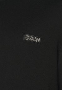 Vêtement à manches longues en maille noire avec une finition texturée et un patch rectangulaire du logo "HUGO" en noir et argent à l'avant.