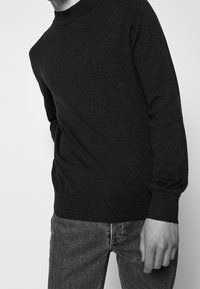 Person in einem dunklen Langarmpullover und abgetragenen Jeans, stehend vor einem schlichten hellen Hintergrund mit einem entspannten Arm.
