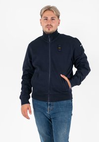 Marine blauwe zip-up sweatshirt met hoge kraag, voorzakken en zwarte hardware details op de borst. Zachte textuur, casual ontwerp.