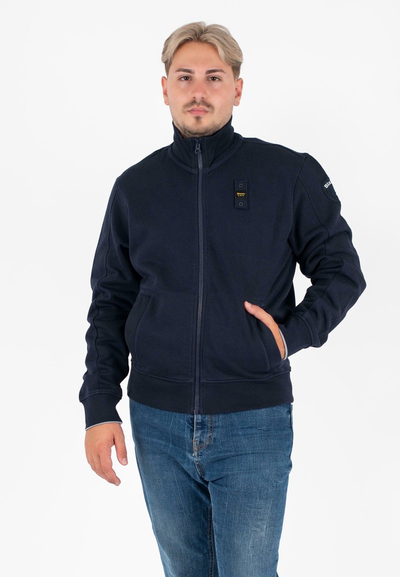 Marine blauwe zip-up sweatshirt met hoge kraag, voorzakken en zwarte hardware details op de borst. Zachte textuur, casual ontwerp.