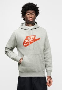 Γκρι φούτερ με κουκούλα και μπροστινή τσέπη, με έντονο κόκκινο λογότυπο Nike «JUST DO IT». Το υλικό φαίνεται μαλακό· το σχέδιο περιλαμβάνει κουκούλα με κορδόνι.
