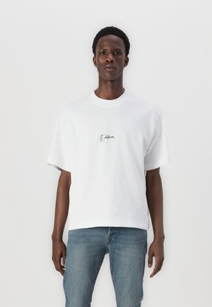 White cotton t-shirt with short sleeves, a crew neck, and black "Edition" text embroidered on the left chest area. Pairing with blue jeans.