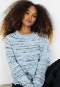 Femeie cu păr voluminos și creț, purtând un pulover tricotat cu dungi albastre deschis și gri, stând pe un fundal deschis.