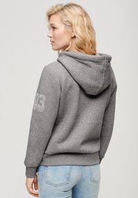 Sudadera gris hecha de una tela suave con un bolsillo canguro y capucha ajustable con cordón. Cuenta con una impresión de logo en la manga izquierda.