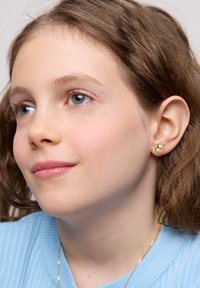 Złote kolczyki wkrętki o gładkim, zaokrąglonym kształcie. Modelka ma jasnobrązowe falowane włosy i nosi jasno-niebieski sweter z prążkowanej tkaniny.